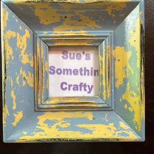 Puede incluir: Un pequeño marco de fotos azul y dorado desgastado con un inserto de papel blanco que dice "Sue's Somethin Crafty".