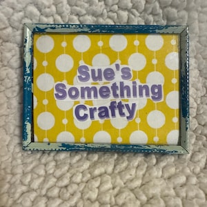 Könnte beinhalten: Ein rechteckiges Schild mit gelbem Hintergrund und weißen Punkten. Das Schild hat einen blauen Rahmen mit einem Used-Look. Der Text "Sue's Something Crafty" ist in lila Buchstaben.