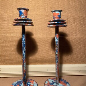 May include: Pair of vintage candle holders. Each holder has a round base, a tall, slender stem, and a top designed to hold a candle. The holders are painted in a distressed style with blue, orange, and white paint.