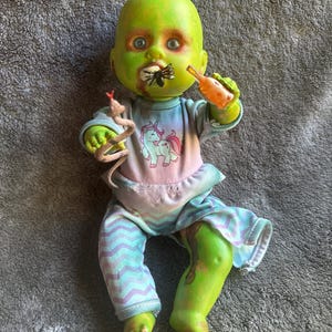 May include: A zombie baby doll with green skin, wearing a pastel unicorn shirt and blue patterned pants. The doll has a spider in its mouth, a small bottle, and a snake. The doll's skin has a decaying appearance.