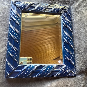 May include: A square mirror with a dark blue, intricately designed frame. The frame features a woven pattern with white accents and small white dots. The mirror reflects a room with wooden paneling.