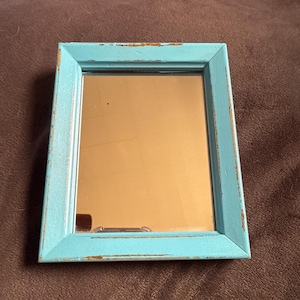 Op de afbeelding: Een kleine, vierkante spiegel met een verweerde, lichtblauwe houten lijst. De lijst heeft een licht verweerde uitstraling, met hints van het onderliggende hout. Het oppervlak van de spiegel reflecteert een warme, gouden tint.