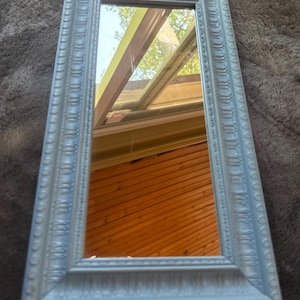 May include: A rectangular mirror with a light blue, ornate frame. The frame has a detailed, textured design. The mirror reflects a view of a wooden surface and a skylight. The mirror is a decorative home decor item.