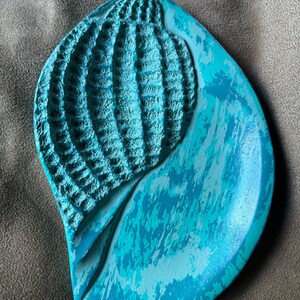 May include: A teal blue ceramic dish with a raised, textured seashell design. The dish has a distressed, painted finish.