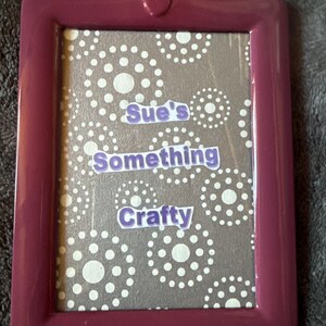 May include: A plum-coloured rectangular picture frame with rounded corners. The frame contains a grey background with white polka dot patterns. The text "Sue's Something Crafty" is displayed in purple lettering.
