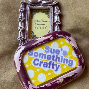 Könnte beinhalten: Zwei violette Bilderrahmen. Der obere Rahmen ist verziert mit einer weißen Schleife und floralen Akzenten. Er trägt ein Etikett mit der Aufschrift "Photo Frame Ornament 2.5" x 3.5". Der untere Rahmen ist abgenutzt und hat einen gelben Hintergrund mit weißen Punkten und einen violetten Aufkleber mit der Aufschrift "Sue's Something Crafty".