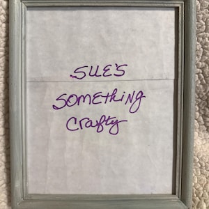 Könnte beinhalten: Ein rechteckiger, grau lackierter Holzbilderrahmen. Der Rahmen enthält ein weißes Papier mit dem handgeschriebenen Text "SUE'S SOMETHING Crafty" in violetter Tinte. Der Rahmen hat ein abgenutztes, verwittertes Aussehen.
