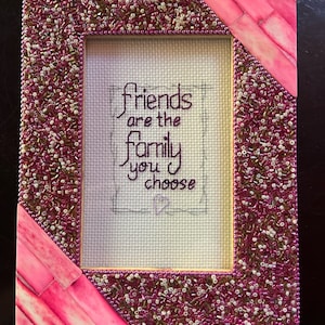Puede incluir: Un marco de fotos de color rosa y blanco con cuentas, con un fondo blanco y el texto "Friends are the family you choose" bordado en hilo negro.