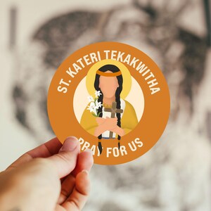 May include: A round sticker featuring an illustration of St. Kateri Tekakwitha. The image shows a woman in a yellow dress holding a cross and flowers, with the text "ST. KATERI TEKAKWITHA" and "PRAY FOR US" in a circular design.