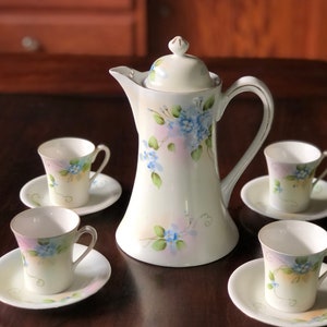 May include: A white porcelain coffee pot with a floral design in shades of blue, pink, and green. The pot has a lid and a handle. There are four matching cups and saucers with the same floral design.