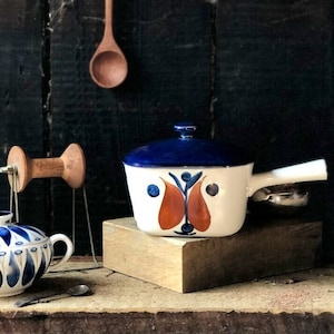 Puede incluir: Una cacerola de cerámica blanca con tapa azul y asa blanca. La cacerola tiene un diseño floral azul y naranja. La cacerola está sobre un bloque de madera.