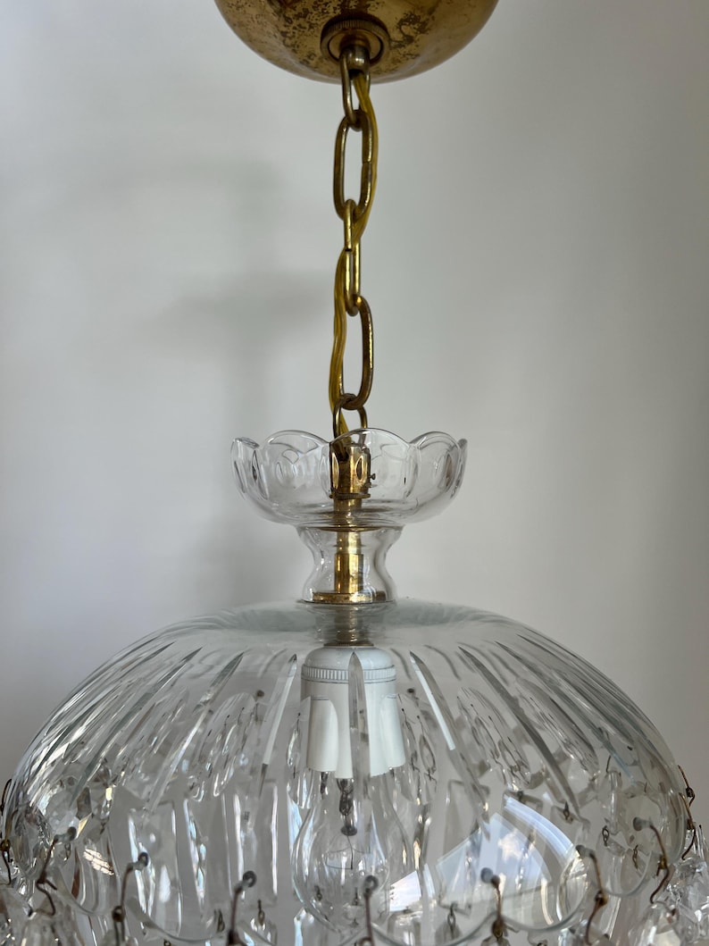 May include: A vintage clear glass chandelier with a gold-colored chain and canopy. The chandelier features a domed shade with vertical ridges and hanging crystal accents. The light fixture has a classic, elegant design.