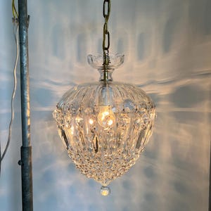May include: A vintage crystal chandelier with a brass-colored canopy and chain. The clear glass shade is adorned with numerous crystal accents and a decorative bottom. The light fixture is suspended from the ceiling, showcasing its intricate design.