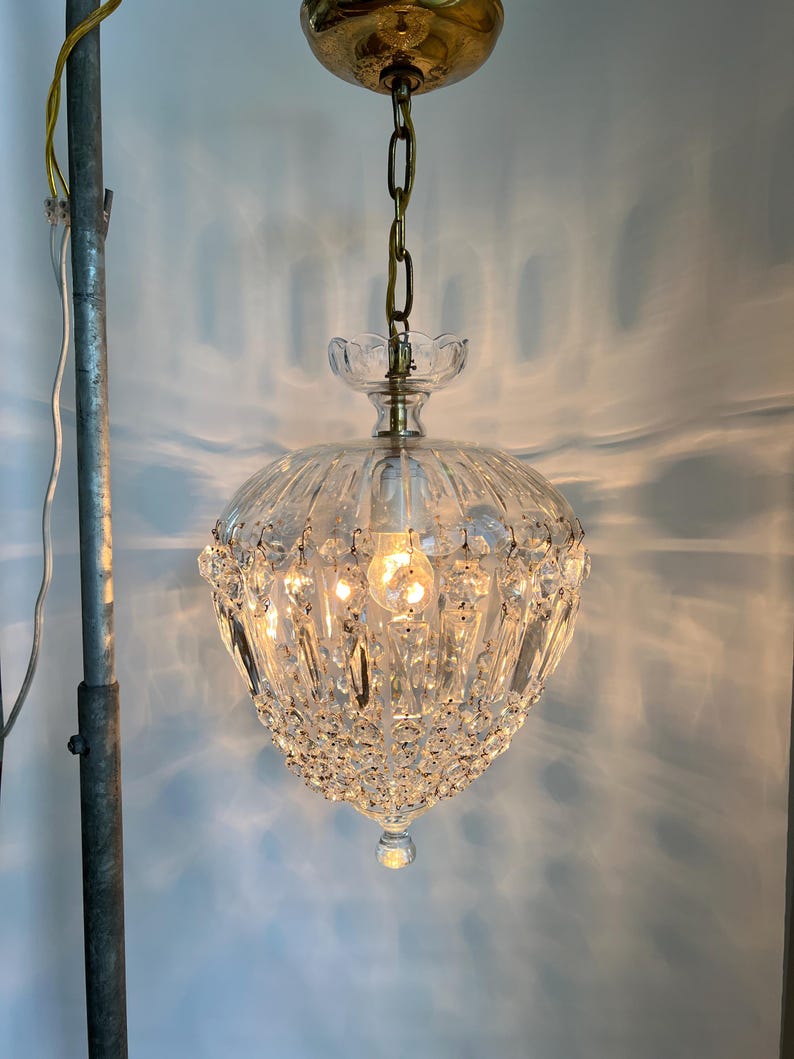 May include: A vintage crystal chandelier with a brass-colored canopy and chain. The clear glass shade is adorned with numerous crystal accents and a decorative bottom. The light fixture is suspended from the ceiling, showcasing its intricate design.
