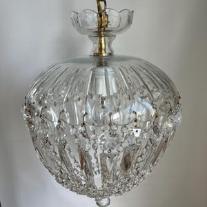 May include: A clear glass chandelier with a round, ribbed shade and hanging crystal accents. The fixture is suspended by a gold chain and features a decorative glass top. The light fixture is a vintage style.