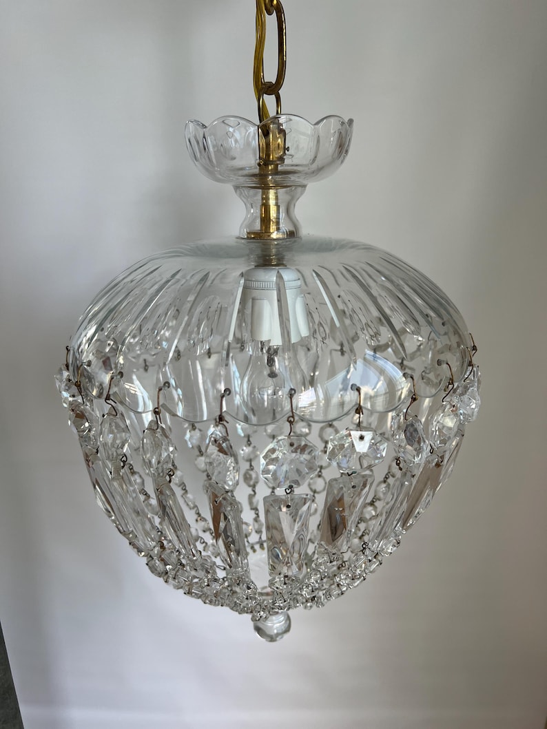 May include: A clear glass chandelier with a round, ribbed shade and hanging crystal accents. The fixture is suspended by a gold chain and features a decorative glass top. The light fixture is a vintage style.