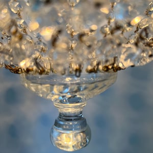May include: Close-up of a clear crystal chandelier with multiple glass pendants. The chandelier has a decorative design with a round base and a series of hanging crystals. The crystals reflect light, creating a sparkling effect.