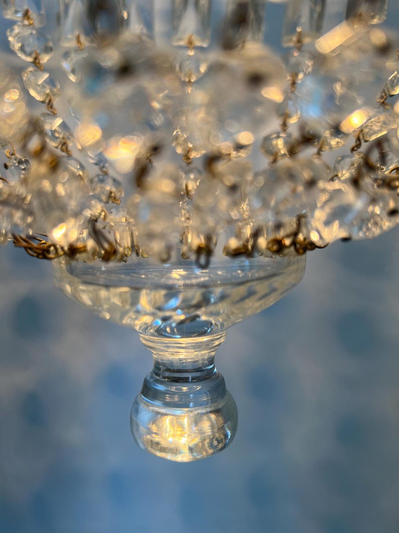 May include: Close-up of a clear crystal chandelier with multiple glass pendants. The chandelier has a decorative design with a round base and a series of hanging crystals. The crystals reflect light, creating a sparkling effect.