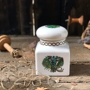 May include: White ceramic jar with a lid featuring a green and brown floral design. The jar has a gold trim around the top and a gold ribbon tied around the floral design.