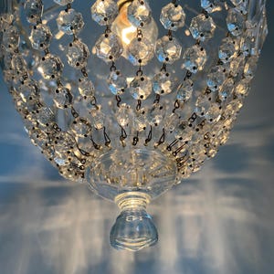 May include: Close-up of a crystal chandelier with multiple faceted crystal beads. The chandelier has a clear glass base and a light bulb in the center. The light casts shadows on the blue wall.