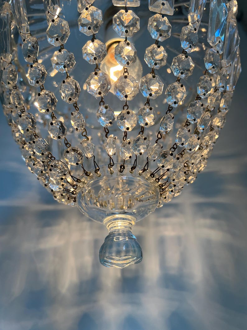 May include: Close-up of a crystal chandelier with multiple faceted crystal beads. The chandelier has a clear glass base and a light bulb in the center. The light casts shadows on the blue wall.