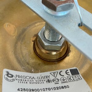 May include: Close-up of a brass-colored light fixture with a metal bracket and a visible nut and bolt assembly. A label with the text "PRECIOSA-LUSTRY" and other technical details is affixed to the fixture.