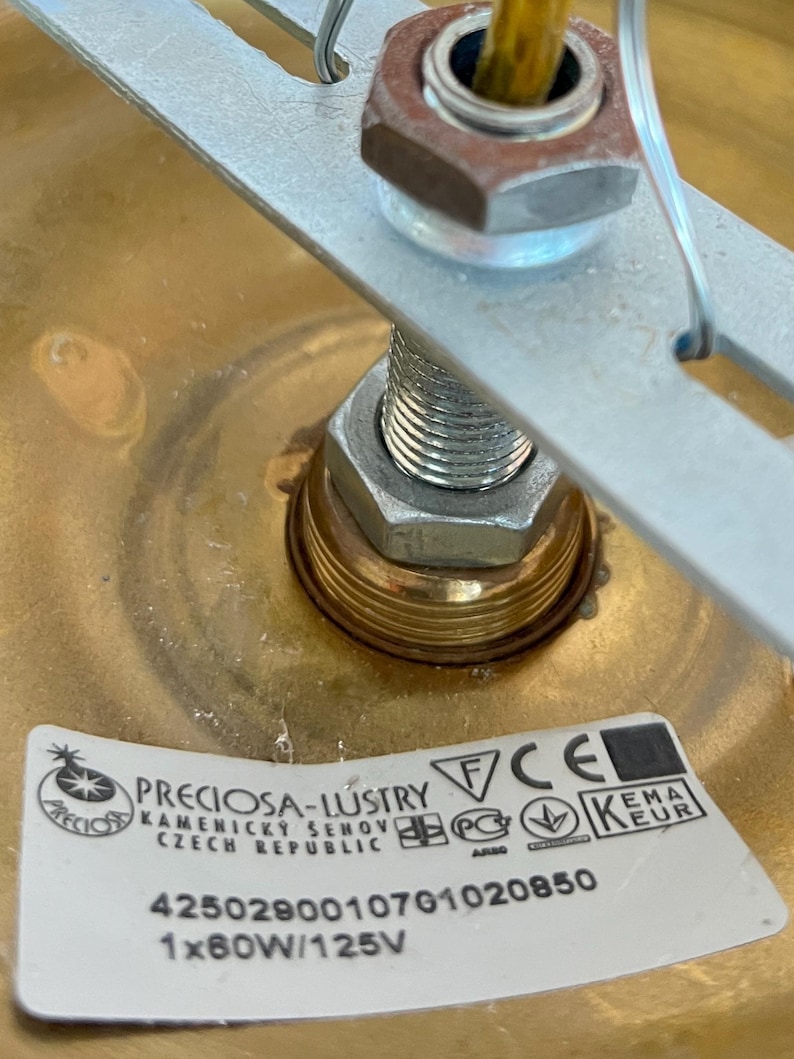 May include: Close-up of a brass-colored light fixture with a metal bracket and a visible nut and bolt assembly. A label with the text "PRECIOSA-LUSTRY" and other technical details is affixed to the fixture.