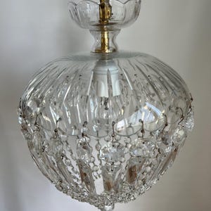 May include: Clear glass chandelier with a round, ribbed shade and hanging crystal accents. The fixture has a brass-colored chain and decorative top. A classic lighting fixture for a dining room or entryway.