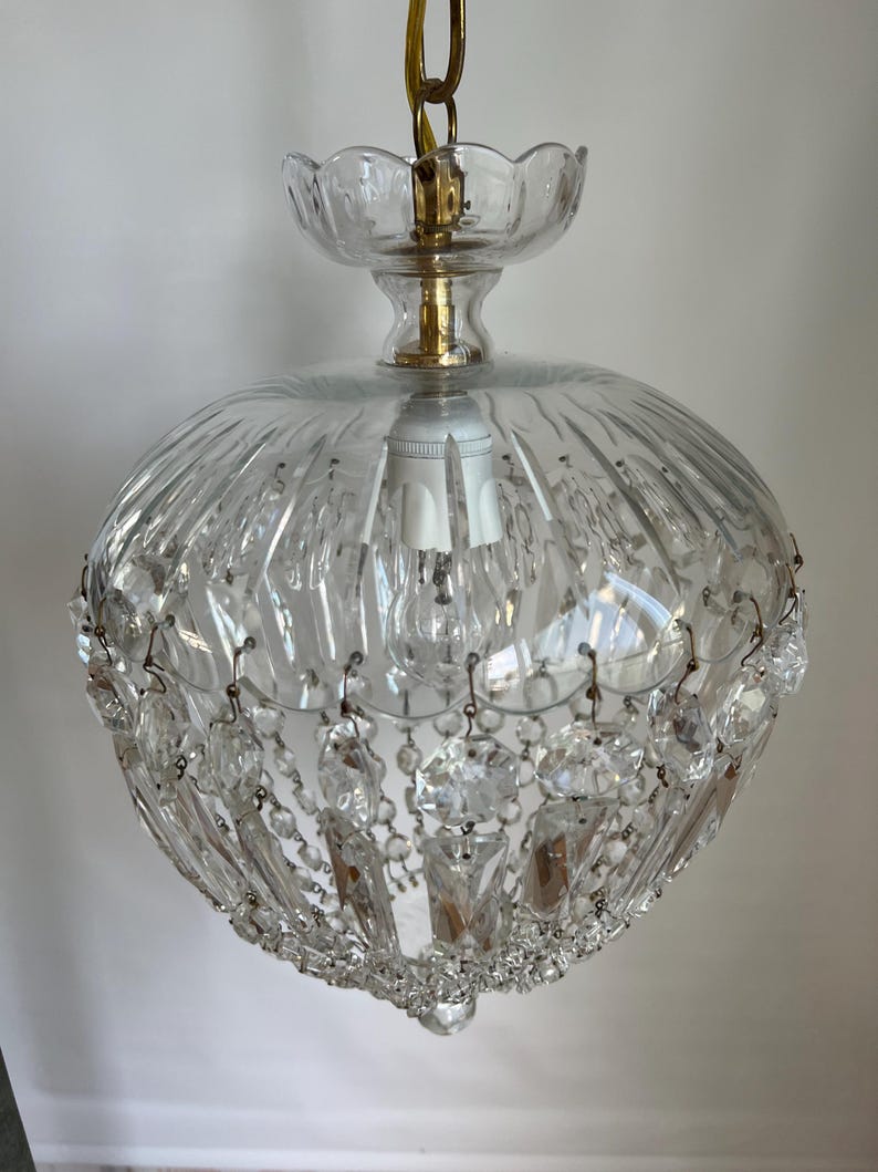 May include: Clear glass chandelier with a round, ribbed shade and hanging crystal accents. The fixture has a brass-colored chain and decorative top. A classic lighting fixture for a dining room or entryway.