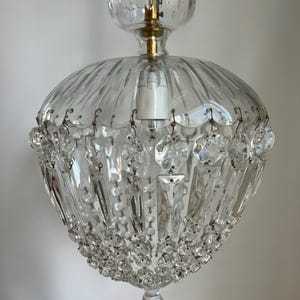 May include: A clear glass chandelier with a scalloped top and a rounded, ribbed body. The chandelier is adorned with hanging crystal prisms and smaller crystal beads. A brass-colored metal fixture holds the lightbulb.