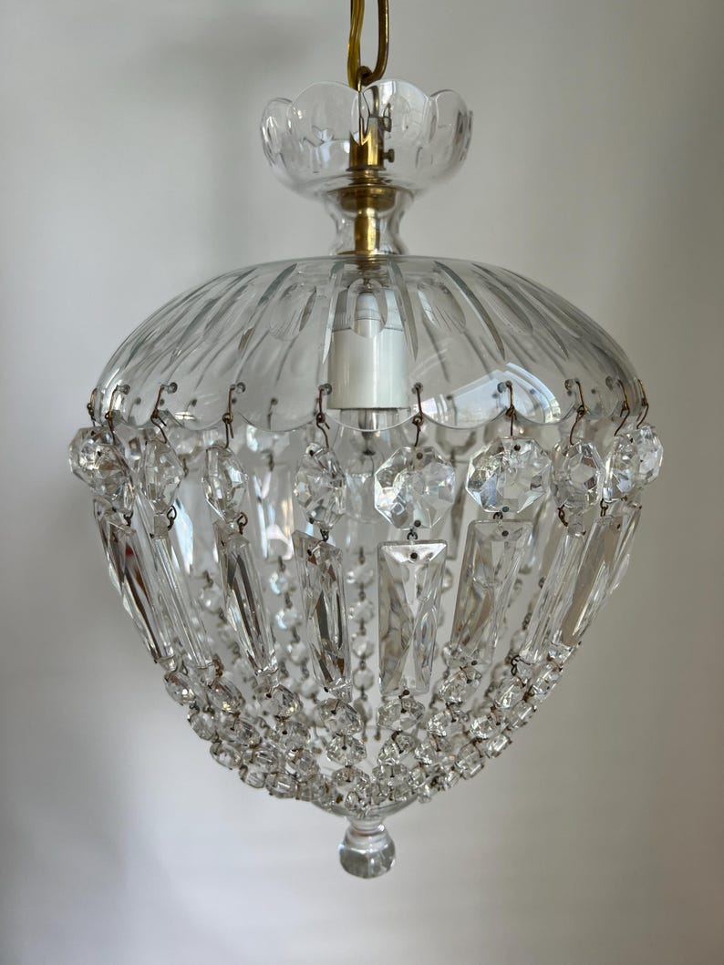 May include: A clear glass chandelier with a scalloped top and a rounded, ribbed body. The chandelier is adorned with hanging crystal prisms and smaller crystal beads. A brass-colored metal fixture holds the lightbulb.
