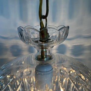 May include: A clear glass pendant light fixture with a decorative, cut-glass shade. The fixture has a scalloped edge and is suspended by a brass chain. The light bulb is visible and lit, casting a warm glow.