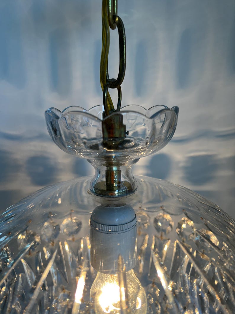 May include: A clear glass pendant light fixture with a decorative, cut-glass shade. The fixture has a scalloped edge and is suspended by a brass chain. The light bulb is visible and lit, casting a warm glow.