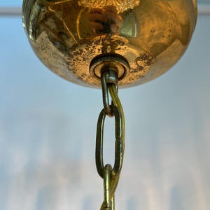 May include: Close-up of a vintage brass ceiling light fixture. The fixture features a rounded, tarnished brass canopy with a chain and a small metal connector. The chain is a gold color.