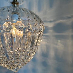 May include: A clear glass chandelier with crystal accents. The light fixture has a rounded shape with a decorative top and hanging crystal pendants. The light is on, casting shadows on the blue wall.
