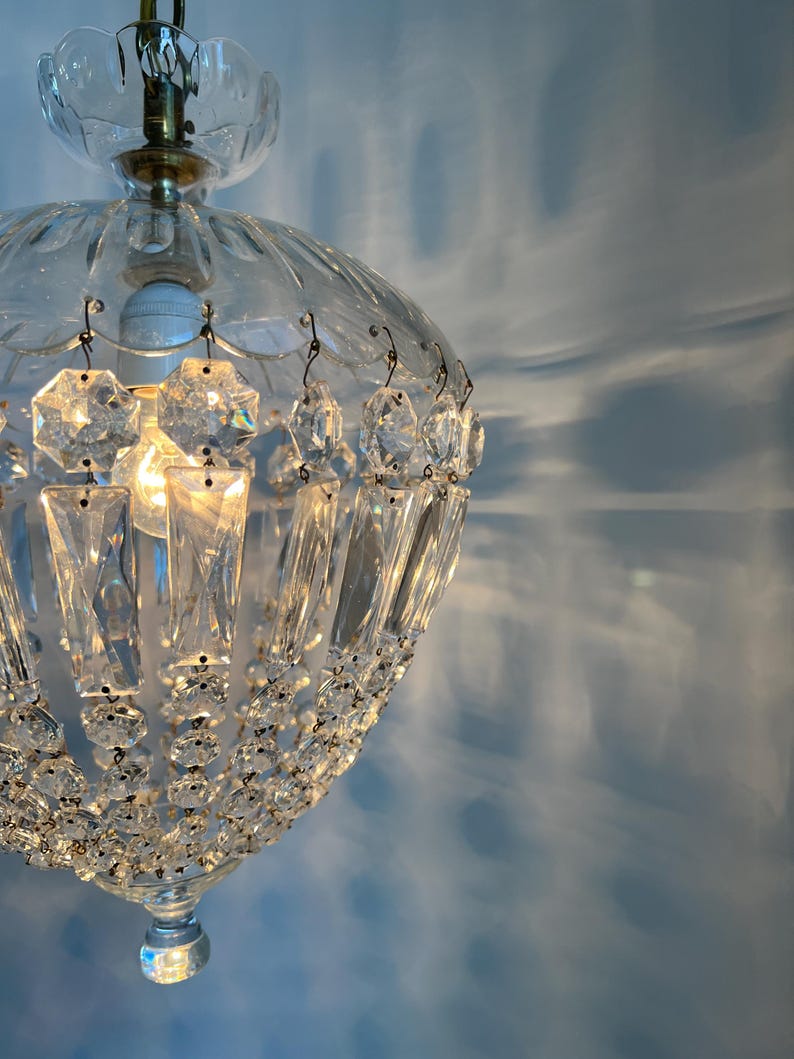 May include: A clear glass chandelier with crystal accents. The light fixture has a rounded shape with a decorative top and hanging crystal pendants. The light is on, casting shadows on the blue wall.