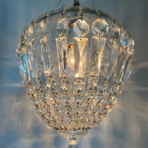 May include: A clear glass chandelier with a round, globe-like shape. The chandelier is adorned with numerous clear crystal accents, including rectangular and octagonal shapes, and small round beads. A light bulb is visible inside.