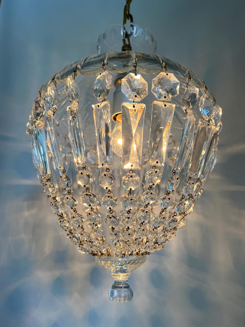 May include: A clear glass chandelier with a round, globe-like shape. The chandelier is adorned with numerous clear crystal accents, including rectangular and octagonal shapes, and small round beads. A light bulb is visible inside.