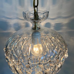 May include: A vintage clear glass chandelier with a brass chain and accents. The light fixture has a round, ribbed glass shade and is adorned with hanging crystal accents. The bulb is lit, casting a warm glow.