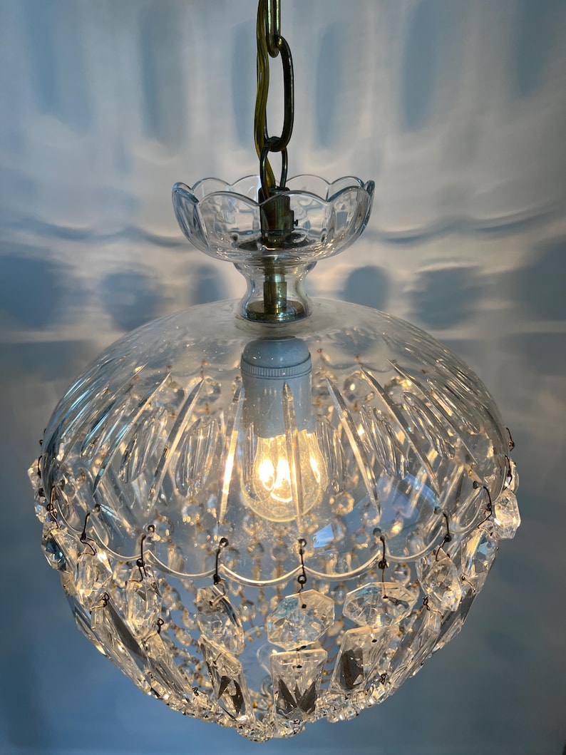 May include: A vintage clear glass chandelier with a brass chain and accents. The light fixture has a round, ribbed glass shade and is adorned with hanging crystal accents. The bulb is lit, casting a warm glow.