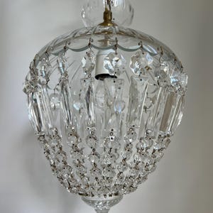 May include: Clear glass chandelier with hanging crystal accents. The light fixture has a rounded shape with a scalloped top and a decorative bottom. The chandelier is suspended by a gold chain.