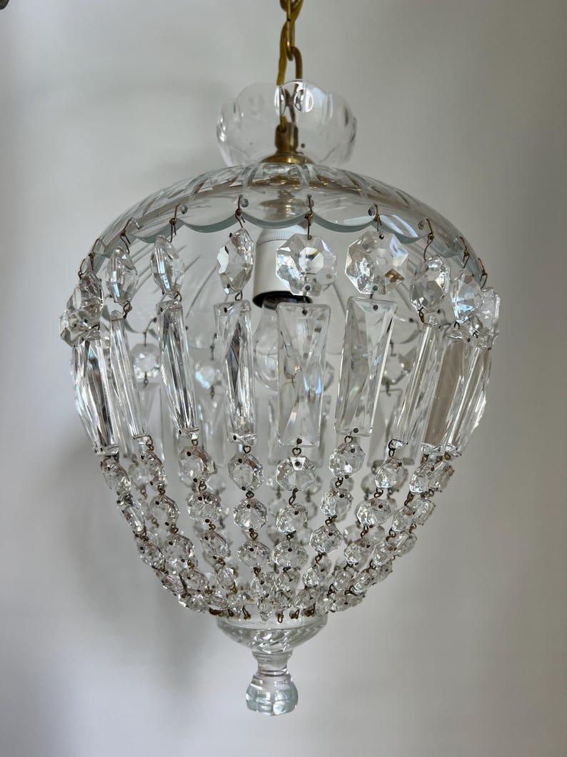 May include: Clear glass chandelier with hanging crystal accents. The light fixture has a rounded shape with a scalloped top and a decorative bottom. The chandelier is suspended by a gold chain.