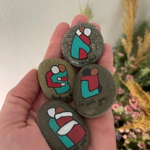 May include: Four painted stones with hugging figures in red, teal, and white. Each stone has a different message: "not alone", "thinking of you", "I'm with you", and "get you". The stones are held in a hand.