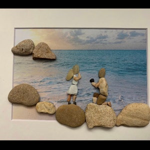 May include: A framed artwork depicting a couple on a beach, created with small stones. The man is kneeling and the woman is standing. The background is a photo of a beach with a sunset.