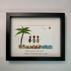 May include: Framed artwork featuring a pebble art design of four figures standing on a beach with a palm tree and the quote "Side by side or miles apart, Sisters are connected heart to heart."