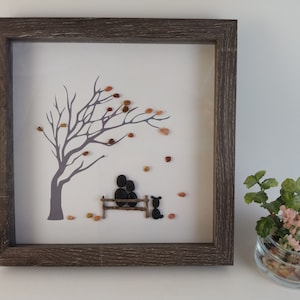 May include: Framed pebble art depicting a couple sitting on a bench under a bare tree with falling leaves. A small dog sits on the ground nearby. The artwork is on a white background with a gray tree and brown pebbles.