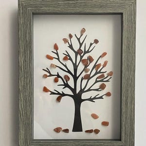 May include: A framed artwork featuring a black silhouette of a tree with small, orange-brown stones arranged to look like leaves. The frame is a light gray wood.