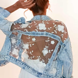 Può includere: Giacca di jeans azzurra con un pannello posteriore trasparente ornato da ricami floreali bianchi. La giacca ha un design classico con bottoni e dettagli di perle. Un capo di abbigliamento esterno elegante e unico.