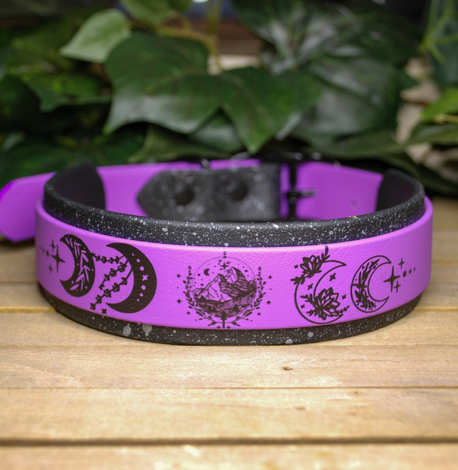 A purple and black cat collar with moon-themed designs on a wooden surface. There are green leaves in the background.