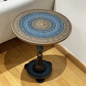 Puede incluir: Una pequeña mesa auxiliar redonda de madera con un intrincado diseño azul y dorado en la parte superior y la base. La mesa tiene una sola pata de pedestal.